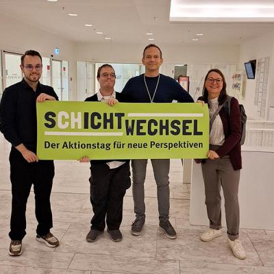 Vier Personen stehen in einem modernen Bürogebäude und halten gemeinsam ein grünes Banner mit der Aufschrift „Schichtwechsel – Der Aktionstag für neue Perspektiven“.