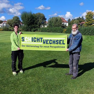 Eine Frau und ein Mann stehen auf einer grünen Wiese am Golfclub und halten ein Banner mit der Aufschrift „Schichtwechsel – Der Aktionstag für neue Perspektiven“