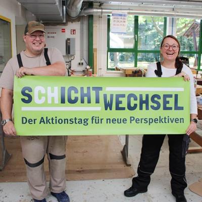 Ein Mann und eine Frau in Arbeitskleidung stehen in einer Werkstatt und halten ein grünes Banner mit der Aufschrift „Schichtwechsel – Der Aktionstag für neue Perspektiven“