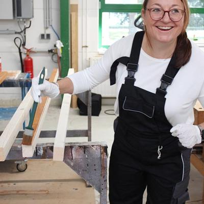 Eine Frau in schwarzer Arbeitslatzhose streicht mit einem Pinsel Holzlatten in einer Werkstatt und lächelt dabei.